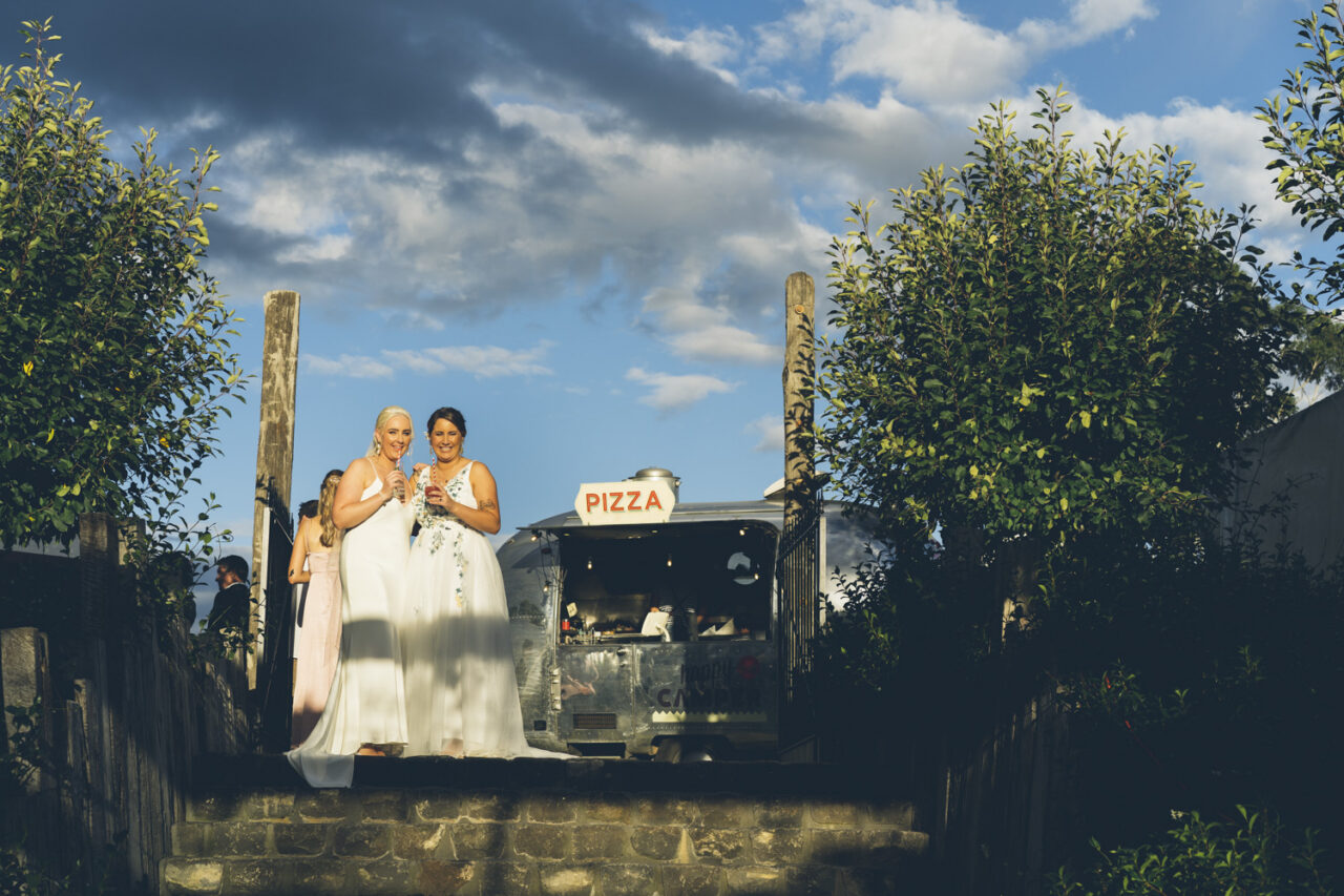 Brides holding drinks near Happy Camper pizza van during wedding reception