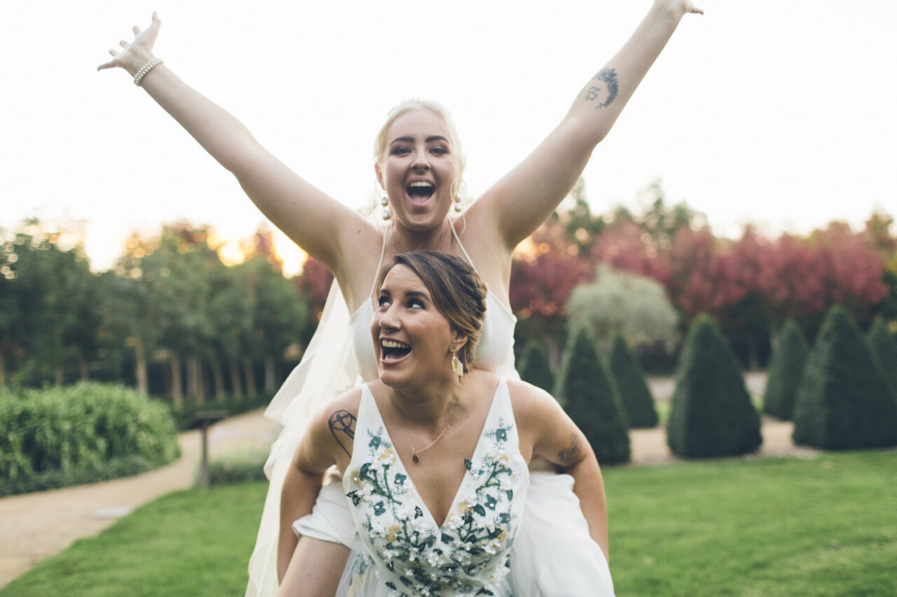 One bride on the other’s back laughing during playful moment at Alowyn Gardens