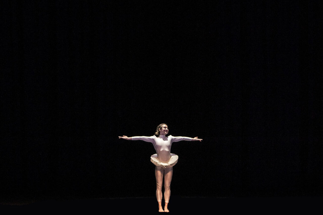 Young ballet dancer in white tutu and leotard performing on a darkened stage with arms outstretched, photographed by LJM Photography