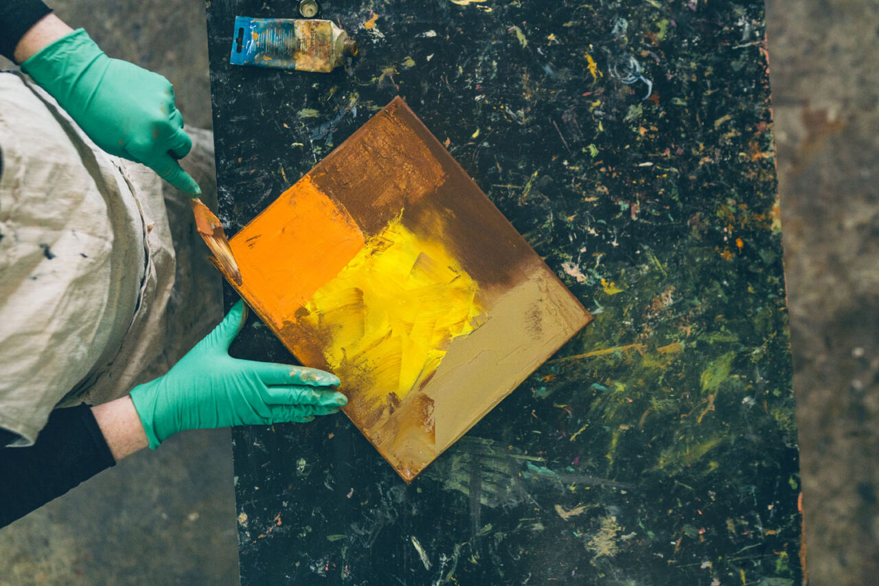 Overhead view of hands in green gloves applying bold yellow and orange paint to a small canvas on a paint-covered studio bench during an artist documentary session on the Surf Coast