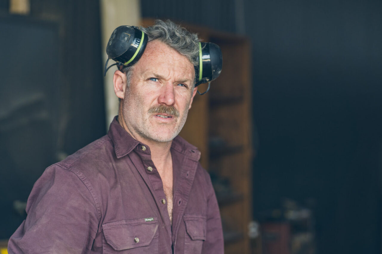 Close up portrait of Anthony Christie wearing ear muffs pushed up on his head, looking directly at the camera in his Ashmore Arts workshop on the Surf Coast