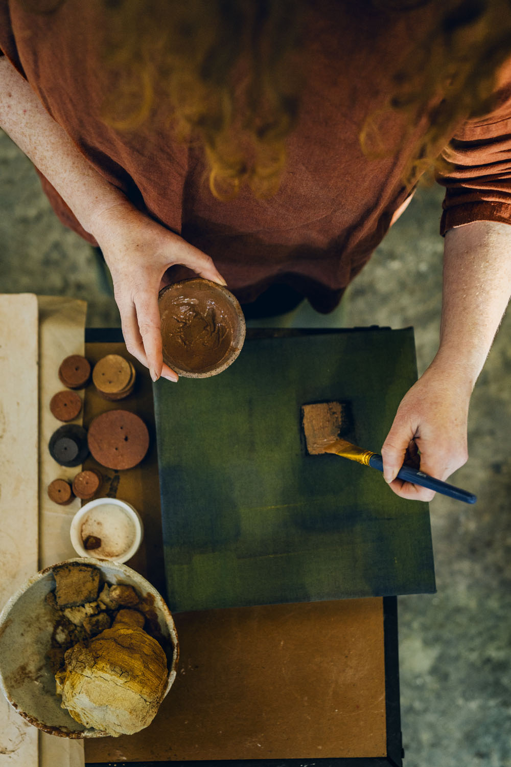 Overhead view of an artist's hands applying clay slip to a dark green ceramic surface with a brush, surrounded by raw clay vessels and pigment pots during a documentary studio session on the Surf Coast