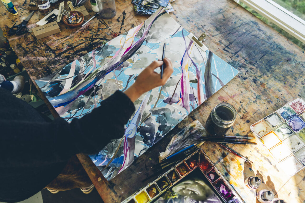 Close up overhead view of an artist's hand painting a large watercolour work on a paint-stained studio desk at Ashmore Arts Torquay, surrounded by brushes, paint palettes and art supplies