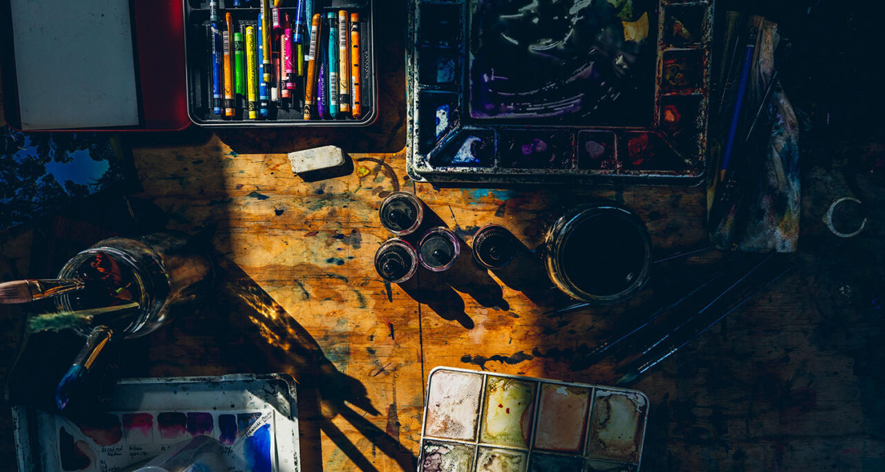 Overhead flat lay of a paint stained artist's desk at Ashmore Arts Torquay showing watercolour palettes, ink jars, pastels, brushes and mixed media supplies bathed in warm light
