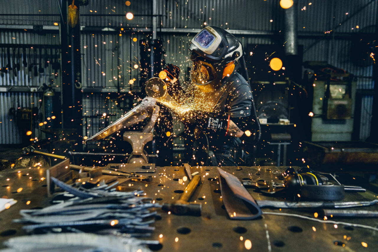 David Graham grinding a metal sculpture with sparks flying across his workshop bench at Ashmore Arts Torquay, wearing full protective gear with blacksmithing tools laid out in front