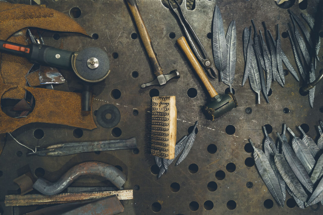 Overhead flat lay of a blacksmith's workbench at Ashmore Arts Torquay showing handcrafted metal feathers alongside hammers, grinders, tongs and wire brushes