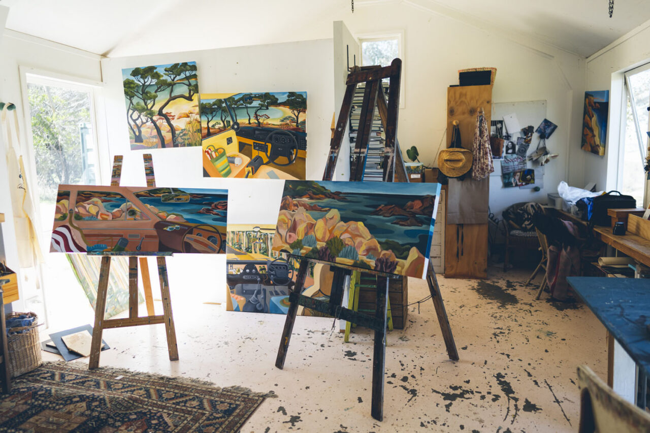 Interior of Kathryn Junor's art studio showing multiple large colourful landscape paintings on easels depicting coastal and bush scenes with a ladder and art materials throughout