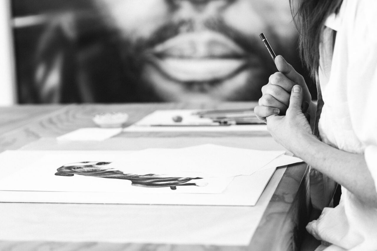 Black and white close up of Kirsten Walsh's hands holding a pencil over a detailed charcoal drawing on her studio desk at Ashmore Arts Torquay, large portrait artwork soft in the background