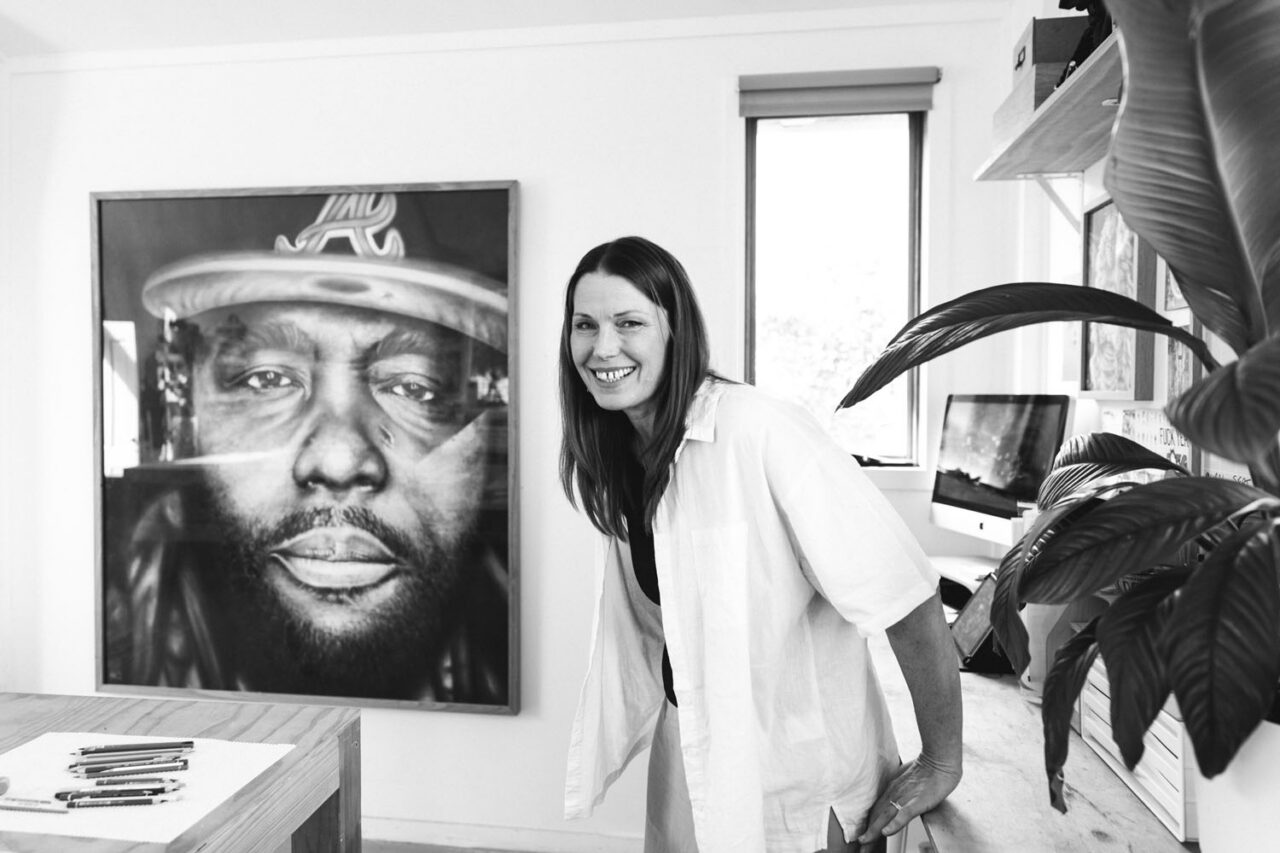 Kirsten Walsh smiling in her Ashmore Arts studio in Torquay, standing beside a large-scale hyper-realistic charcoal portrait artwork