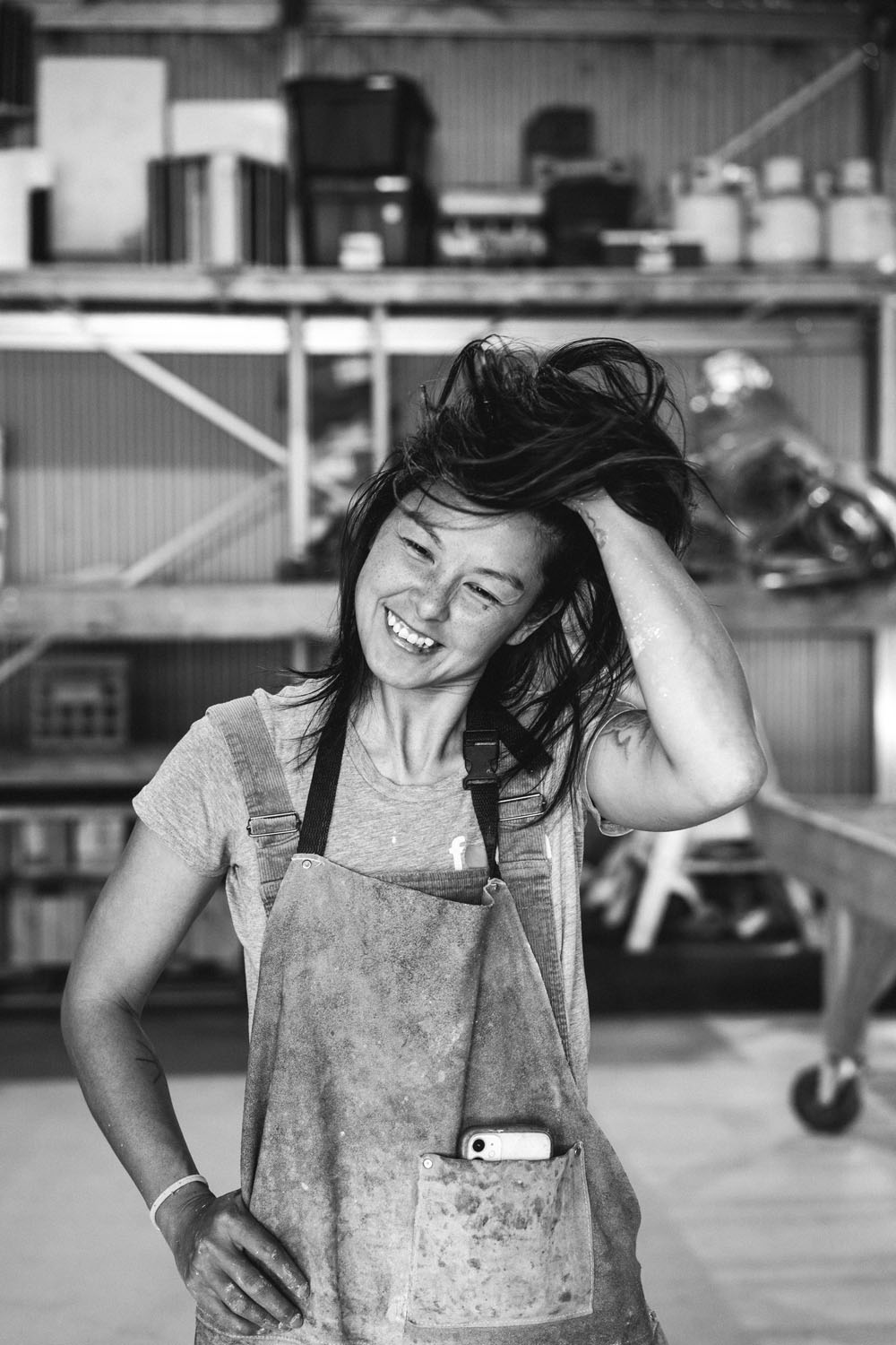 Black and white portrait of Mayde Concrete artist laughing and running her hand through her hair in her workshop at Ashmore Arts Torquay, wearing paint-stained overalls