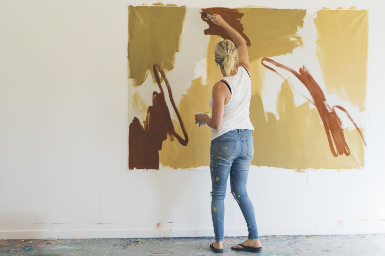 Rowena Martinich seen from behind painting a large earth-toned abstract canvas mounted on a white wall in her Surf Coast studio, wearing paint-splattered jeans