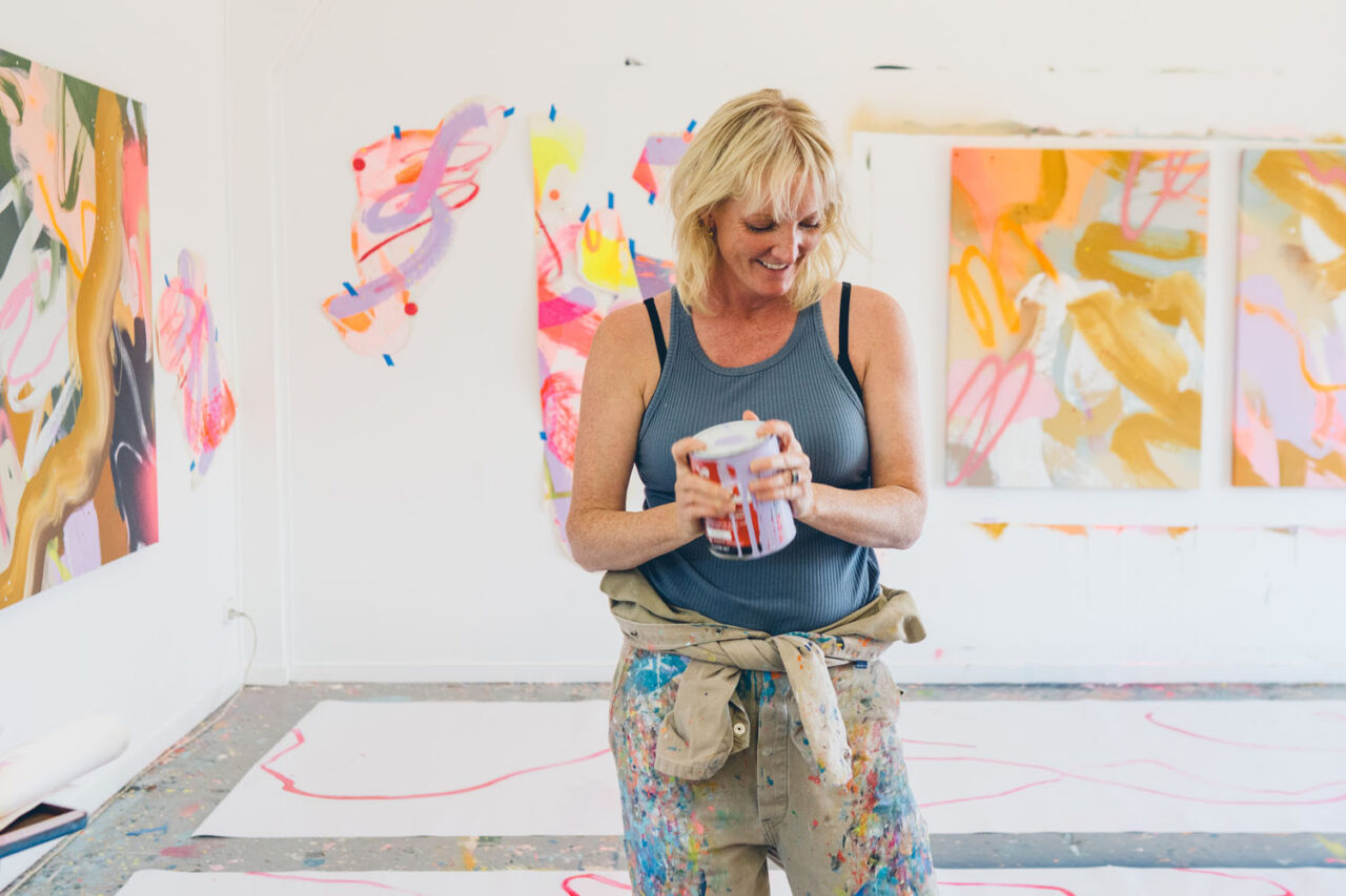 Rowena Martinich standing in her Ashmore Arts studio in Torquay surrounded by large bold colourful abstract paintings, holding a paint tin and smiling