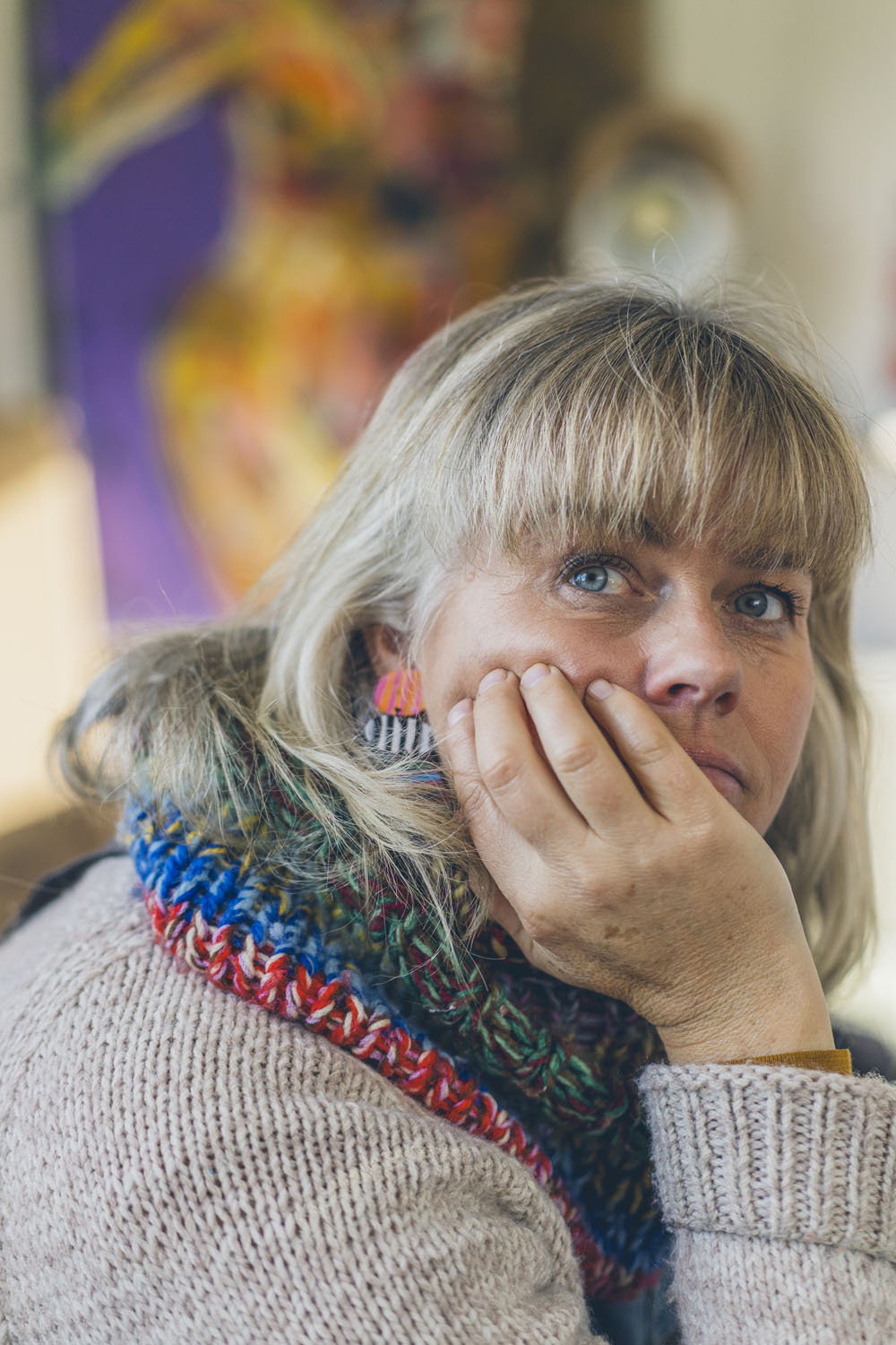Close up portrait of Sam Leggett resting her chin on her hand with a thoughtful expression, colourful artwork softly out of focus in the background