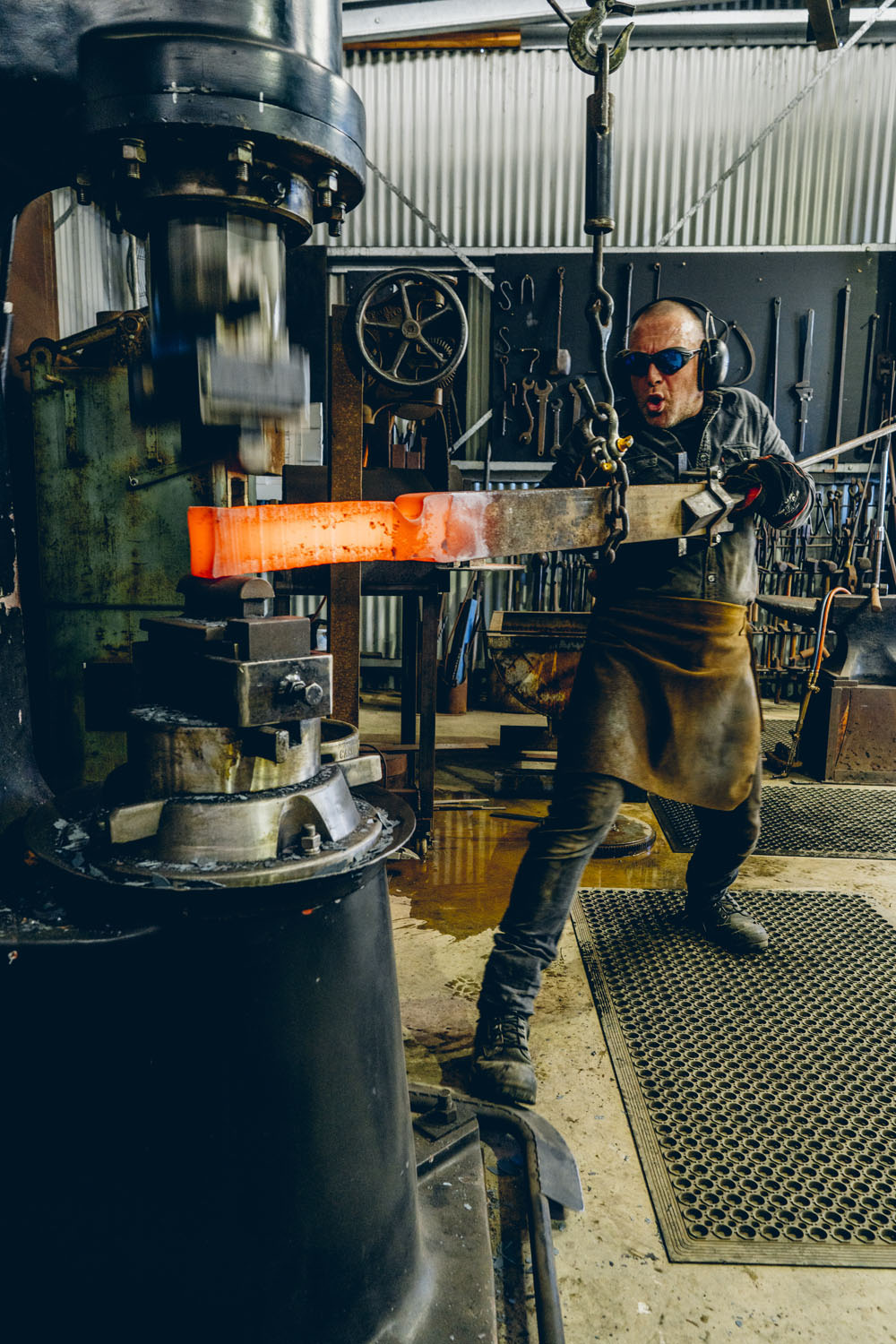 Blacksmith working with glowing orange hot metal on a power hammer in the Simply Forged workshop at Ashmore Arts Torquay, wearing protective gear with tools hanging on the wall behind