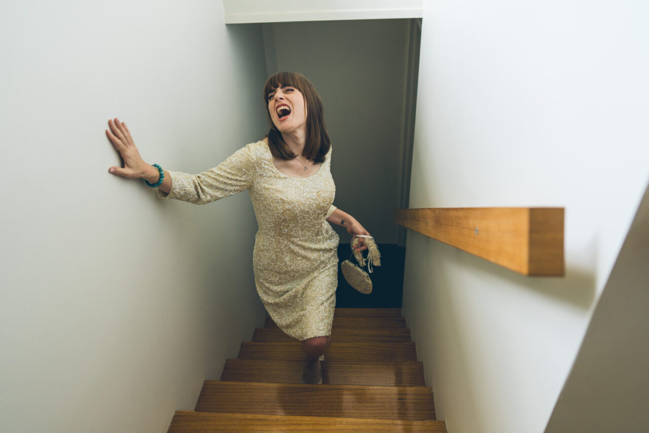 Bride walking up the stairs laughing and holding her shoes during candid pre-wedding moments for Bel and Leigh’s wedding.