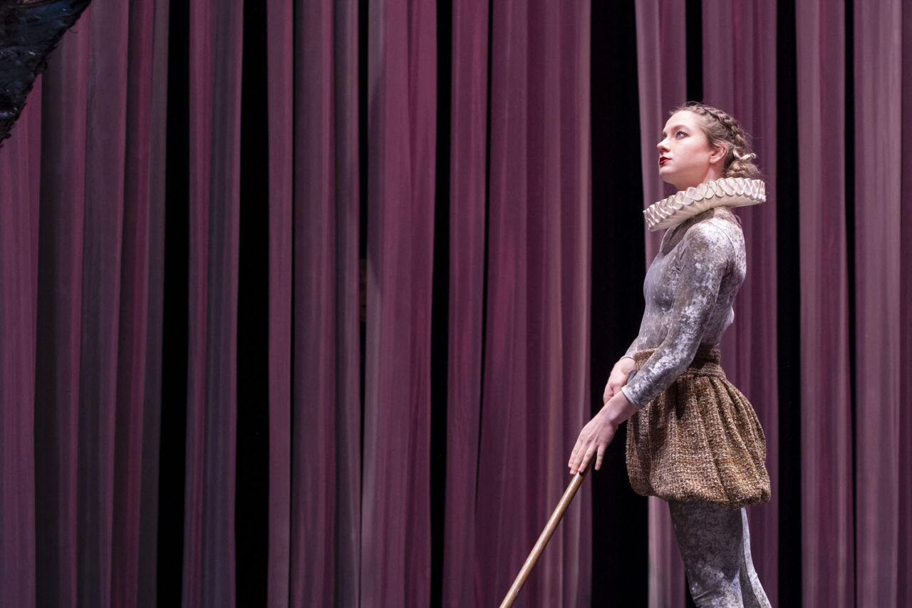 Performer from Circa’s Duck Pond stands centre stage at The Round Melbourne, wearing a ruffled collar and gold costume, lit by soft theatre lighting and mauve curtains.
