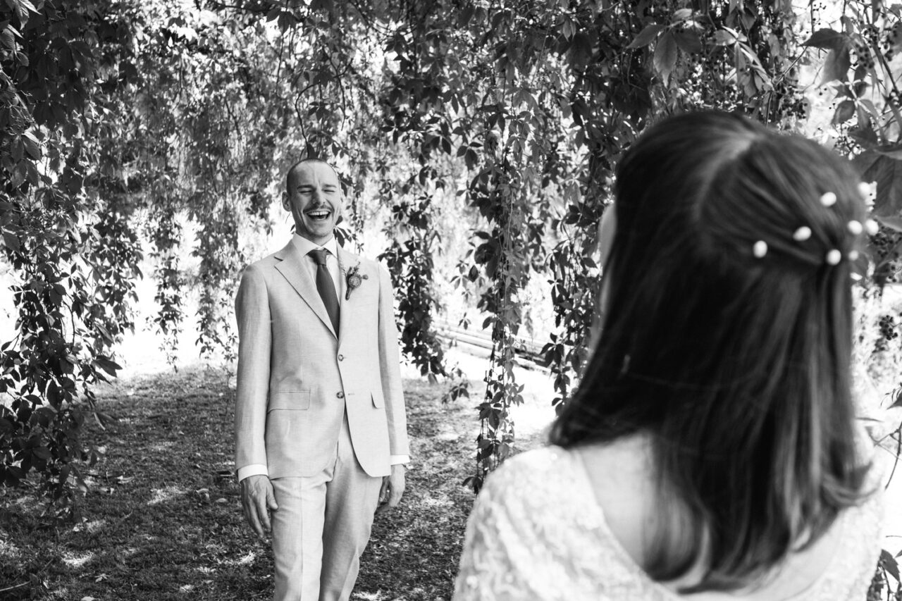 Groom laughing joyfully during first look as he sees his bride under the willow tree canopy.