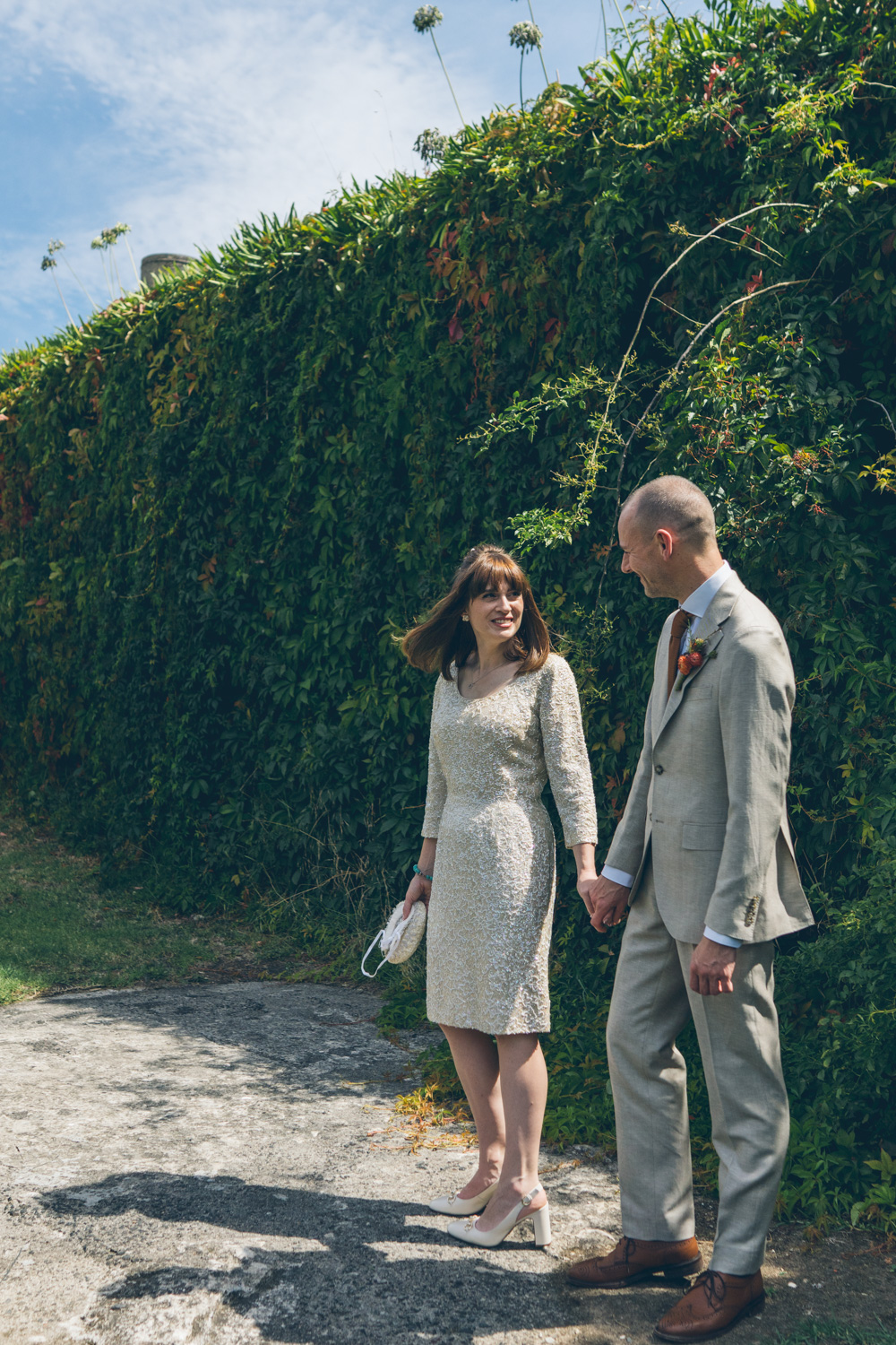 Bride and groom walking hand in hand beside ivy-covered wall, smiling under blue sky