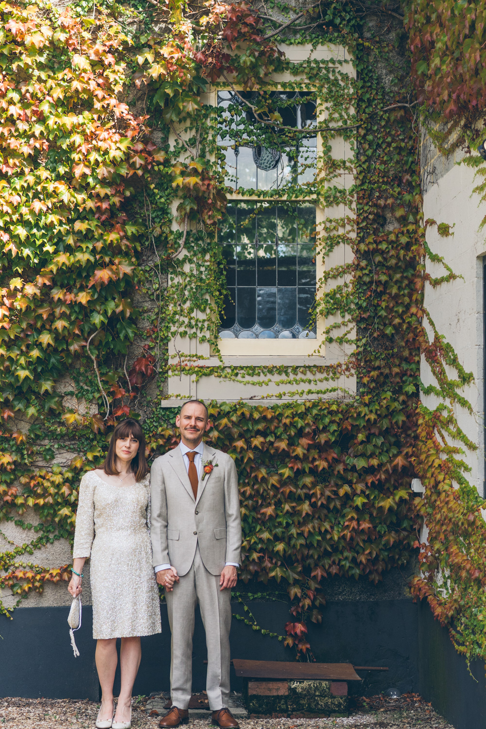 Wedding couple holding hands standing under ivy-covered window outside venue