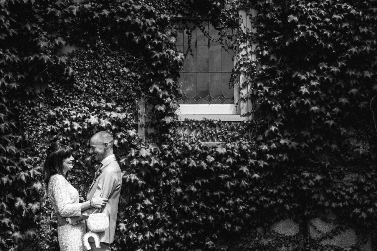 Black and white wedding portrait of couple smiling and embracing under ivy-covered window