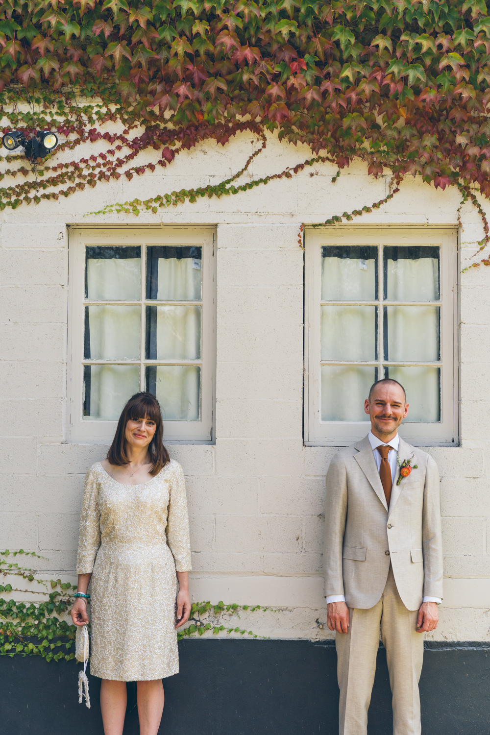 Bride and groom standing apart against white brick wall with twin windows and ivy creeping above