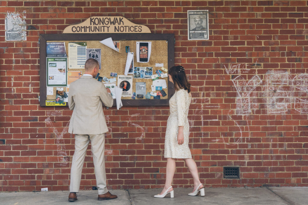 Bride and groom reading community notices on brick wall outside Kongwak Hall