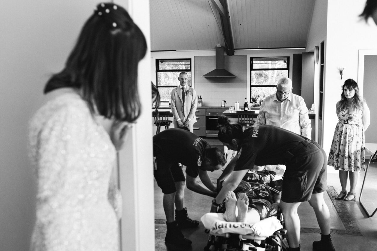 Documentary wedding photo showing paramedics assisting a guest while the bride and groom watch with concern inside the venue.
