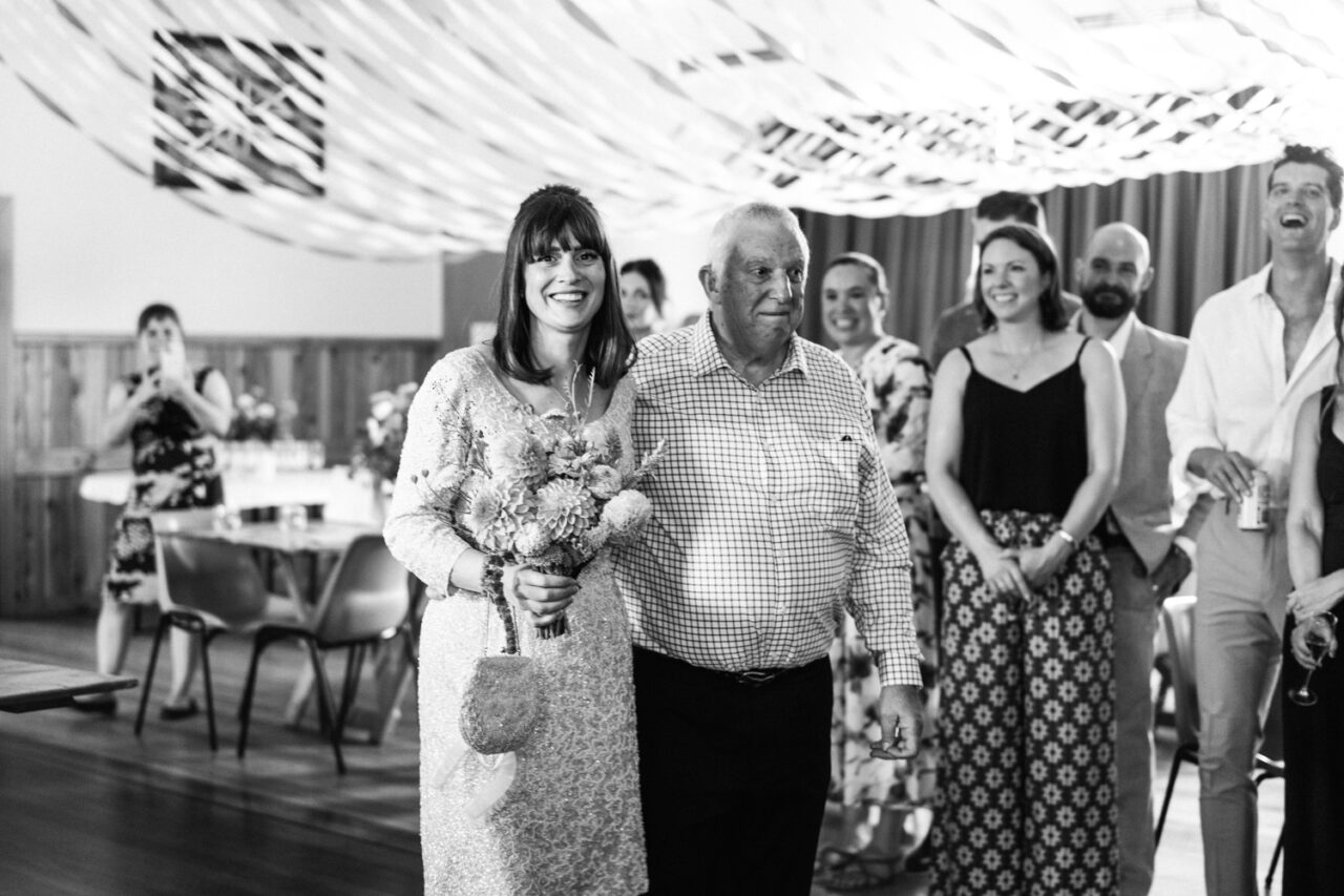 Bride walking down the aisle with her father, smiling at guests gathered in the hall.
