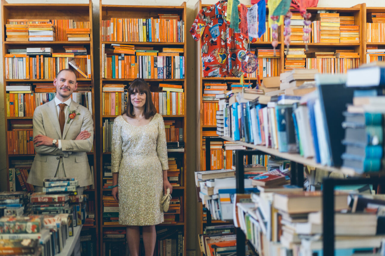 Bride and groom stand together in front of colourful bookshelves, their expressions calm and content in a cosy vintage bookshop.