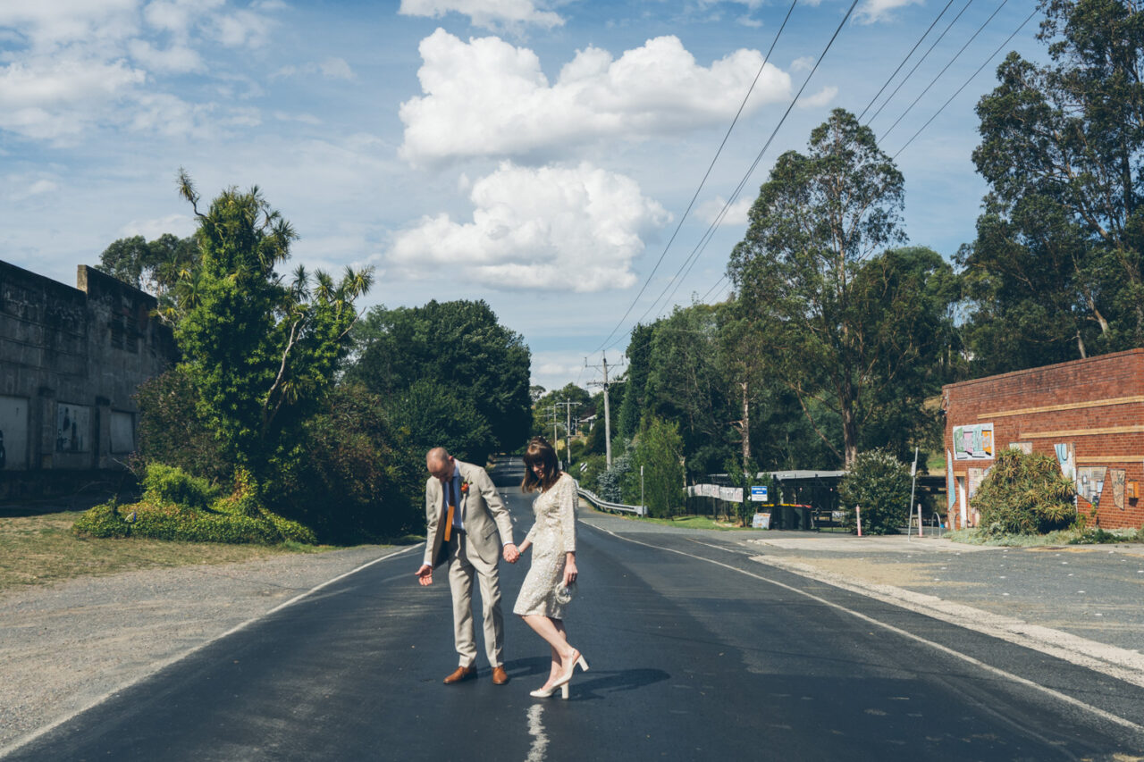Bride and groom walk hand in hand down a quiet country road, sharing a lighthearted moment beneath a blue sky.