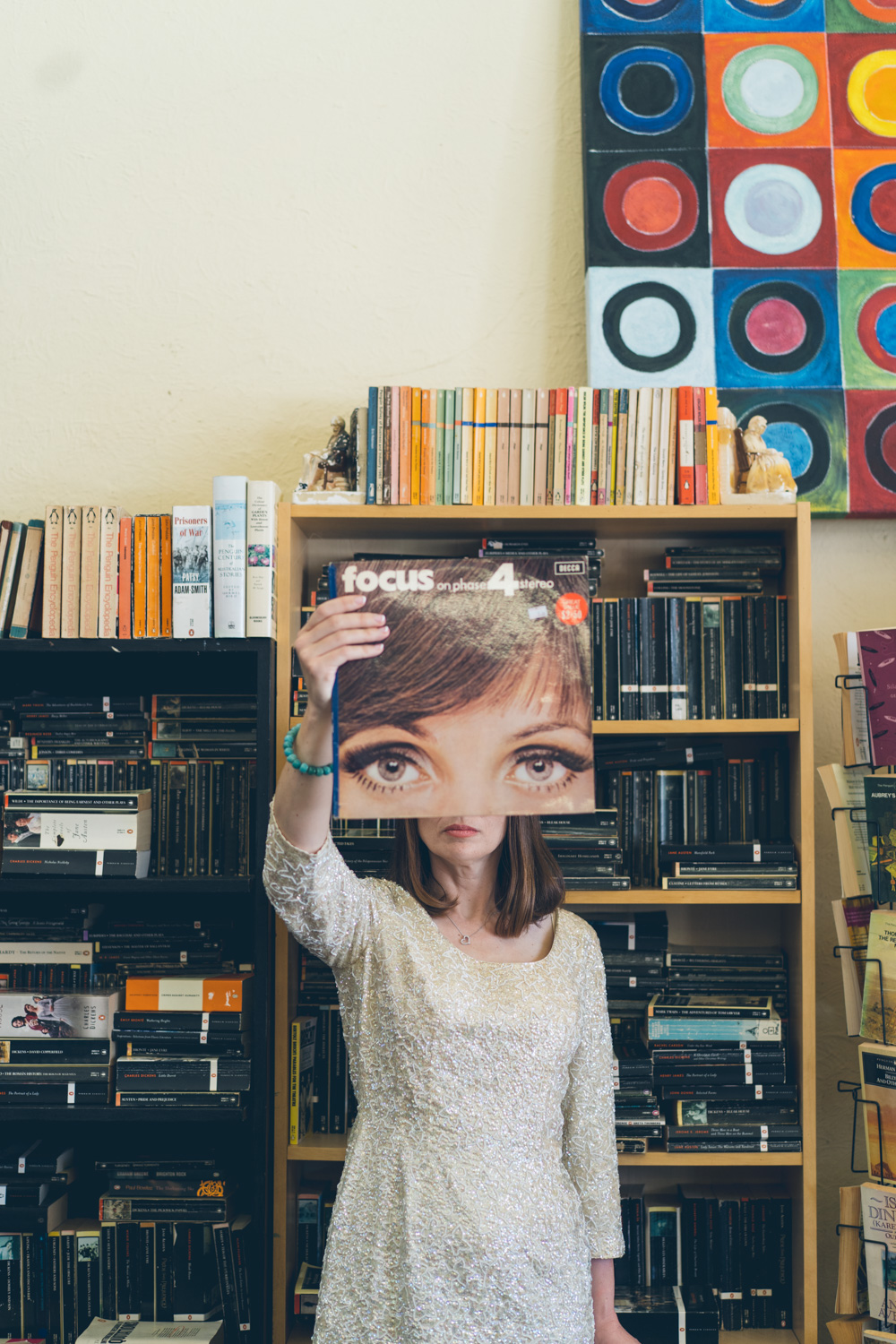 Bride holds up a vintage album cover with large eyes, creating a playful illusion in front of a bookshelf filled with records and books.
