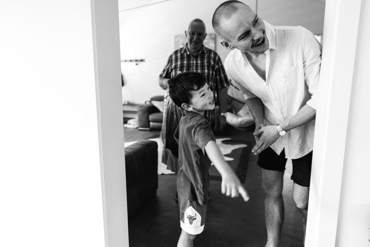 Candid black and white moment of the groom laughing with family and a young boy during morning preparations before Bel and Leigh’s wedding.