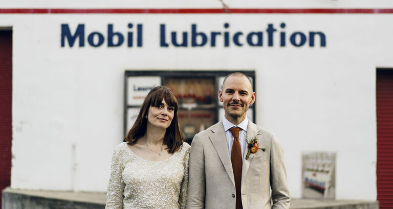 Bride and groom, Bel and Leigh, standing in front of an old Mobil Lubrication building during their candid wedding portrait session.