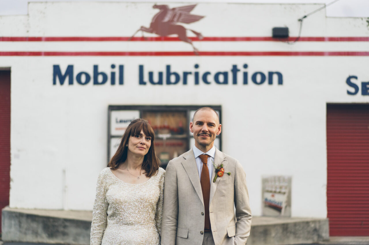 Bride and groom standing outside Mobil Lubrication service station wall