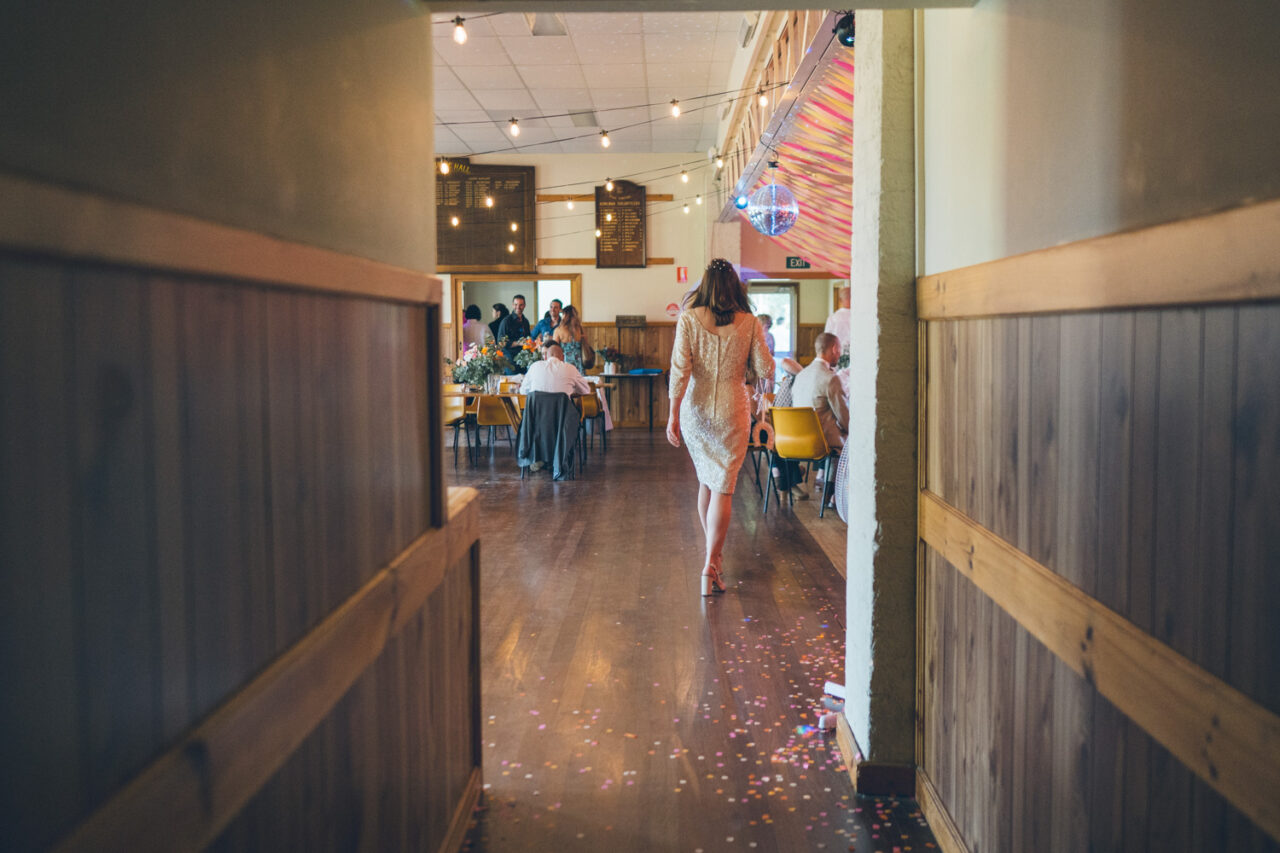 Bride walking down a wooden hallway towards the reception filled with guests and confetti.