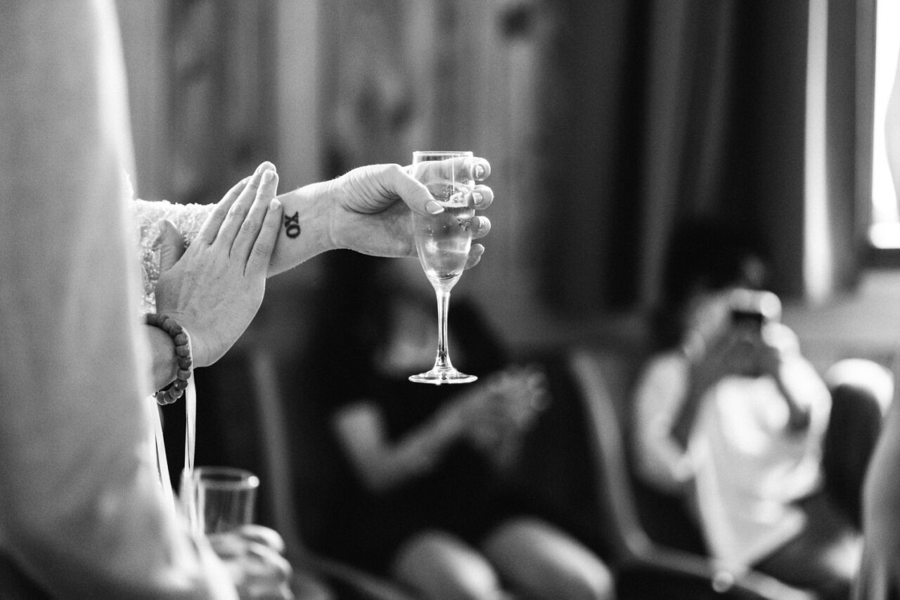 Close-up of champagne glass raised in a toast, showing small XO tattoo on wrist.