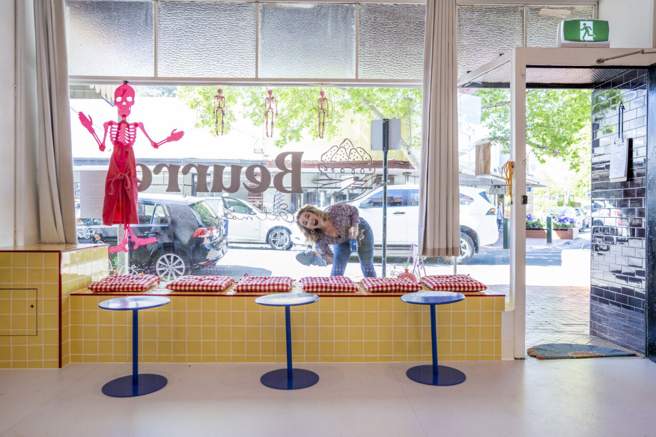 Bek from Kestrel Lane smiling through the front window of Beurre Cakes while cleaning the glass, captured as documentary brand photography for a creative small business.