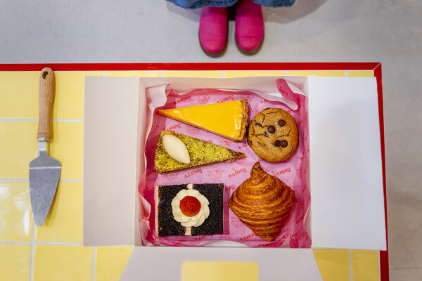 Open pastry box on a yellow tiled counter at Beurre Cakes showing a selection of cakes, a cookie and a croissant, photographed as documentary brand photography for a creative small business.
