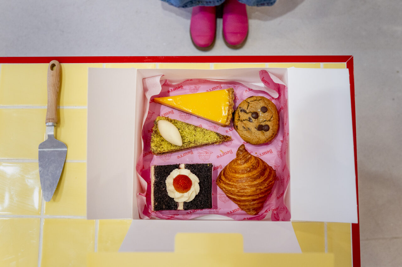 Top down view of a Beurre Cakes pastry box filled with colourful treats on a yellow tiled counter, photographed during documentary brand photography.