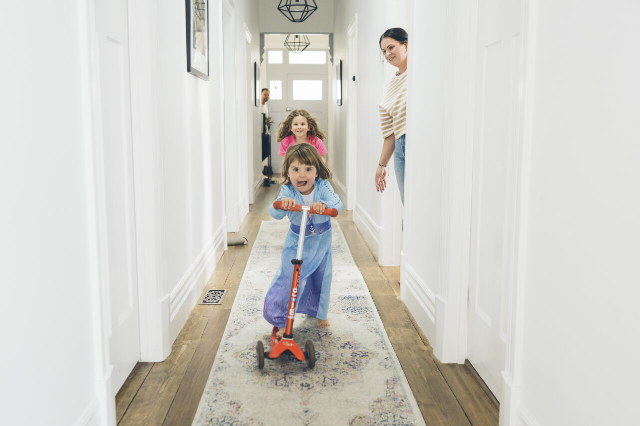 Children racing scooters down the hallway at home while parents watch during a candid summer family photo session Surf Coast
