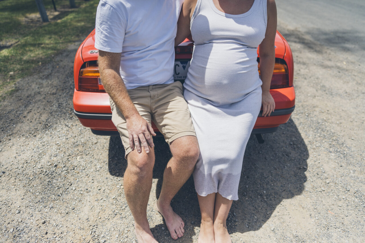 Expecting couple sitting barefoot on the back of their car in the summer sun during a candid photo session on the Surf Coast