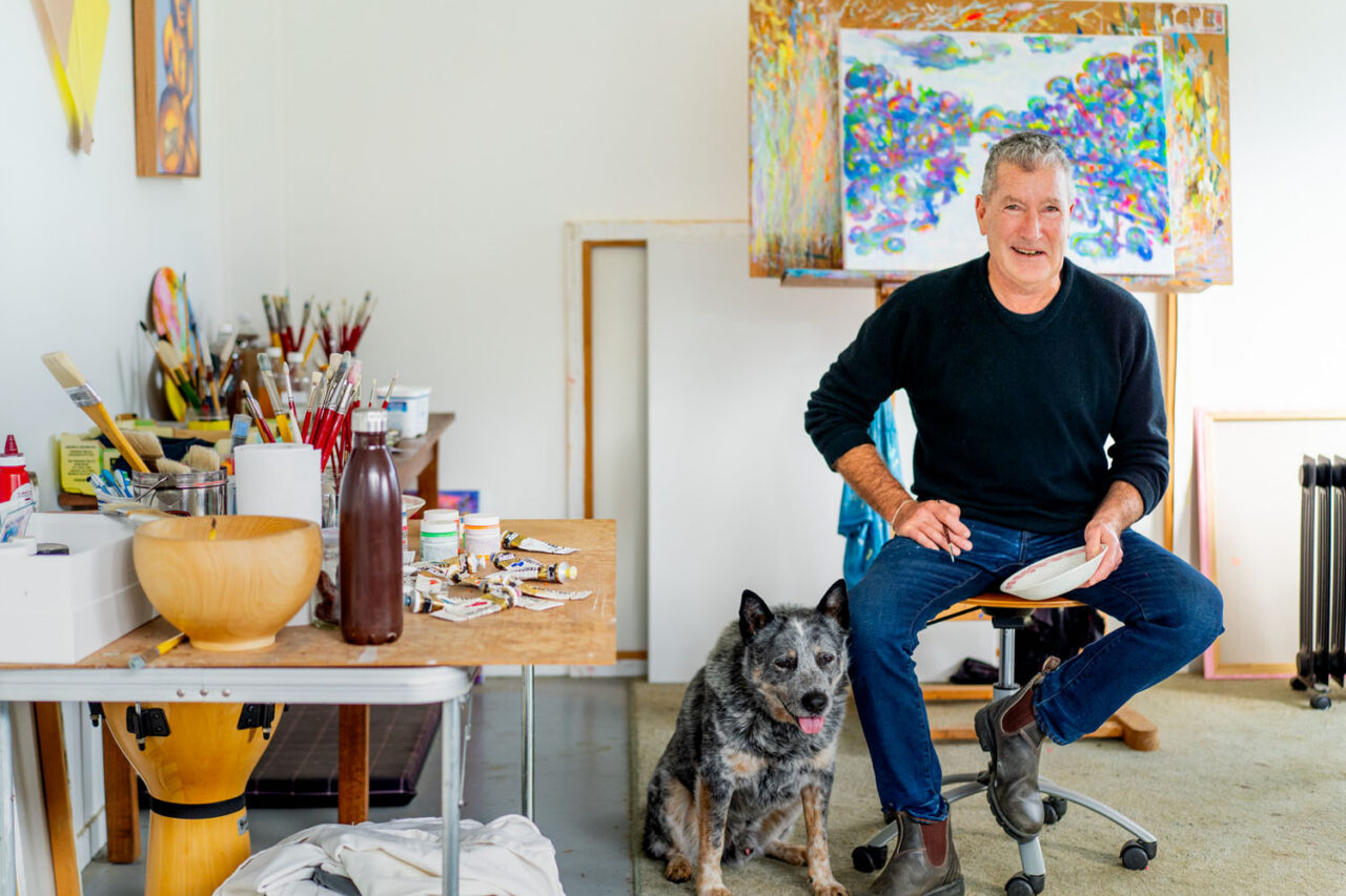 David Rossiter sitting in his Ashmore Arts studio with his dog, surrounded by paint and brushes