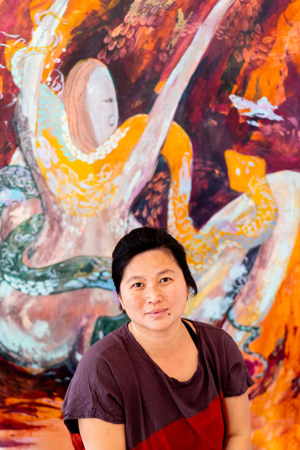 Close up portrait of artist Lily Yap standing in front of her vibrant painting at Ashmore Arts