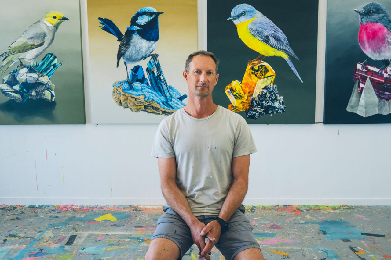 Artist Geoffrey sitting in his Ashmore Arts studio surrounded by detailed bird paintings