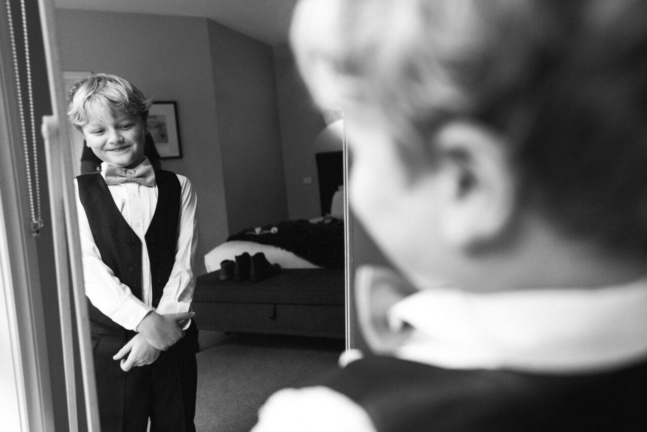 Child in a bowtie smiling at their reflection while getting ready for a wedding, candid family moment