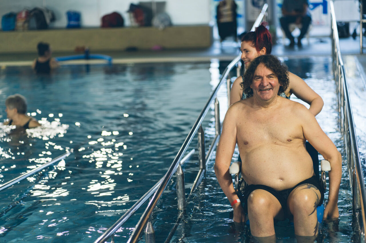 YMCA member using pool access chair with support staff in aquatic centre, documentary lifestyle brand photography.