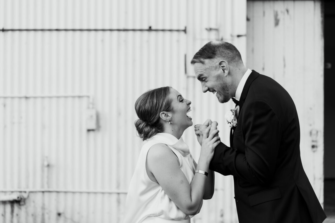 Couple laughing together in front of an industrial wall, relaxed candid wedding photography