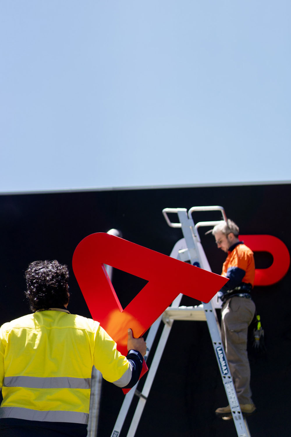 Worker installing large red signage using a ladder, documentary brand photography of industrial installation.