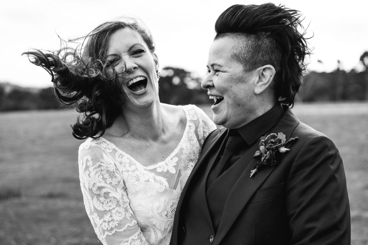 Two brides laughing together outdoors during their wedding portraits.