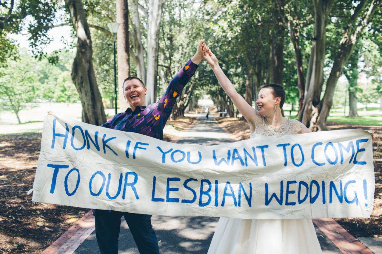 Two brides holding a banner that reads “Honk if you want to come to our lesbian wedding”.